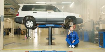 Mechanic in blue coverall using smartphone beside vehicle on lift in repair workshop.