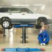 Mechanic in blue coverall using smartphone beside vehicle on lift in repair workshop.