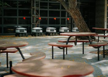 Futuristic delivery robots lined up near outdoor cafeteria tables, showcasing modern technology.