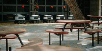Futuristic delivery robots lined up near outdoor cafeteria tables, showcasing modern technology.