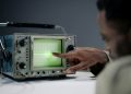 Close-up of a person pointing at a vintage oscilloscope screen in a tech setting.