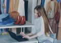 Young woman managing a fashion boutique, multitasking with phone and laptop, surrounded by clothing.