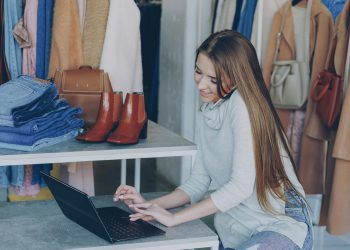 Young woman managing a fashion boutique, multitasking with phone and laptop, surrounded by clothing.