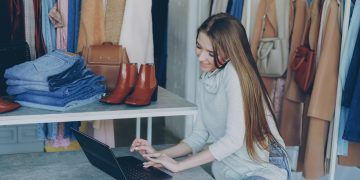 Young woman managing a fashion boutique, multitasking with phone and laptop, surrounded by clothing.