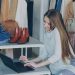 Young woman managing a fashion boutique, multitasking with phone and laptop, surrounded by clothing.