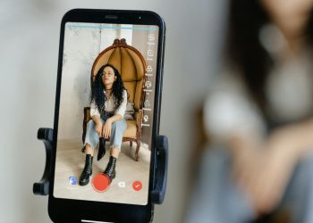 A woman sits in a chair recording a video on her smartphone, showcasing modern vlogging techniques.