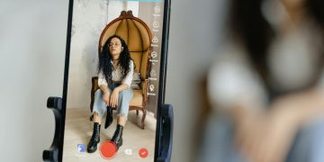 A woman sits in a chair recording a video on her smartphone, showcasing modern vlogging techniques.