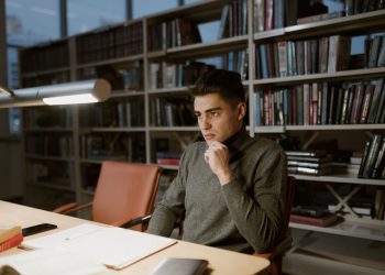 A man in a turtleneck sweater concentrates on studying in a library setting.