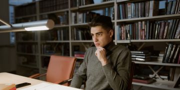 A man in a turtleneck sweater concentrates on studying in a library setting.