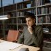 A man in a turtleneck sweater concentrates on studying in a library setting.