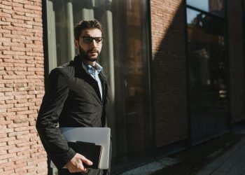 Stylish man in black jacket with glasses holding a laptop outside a glass building.