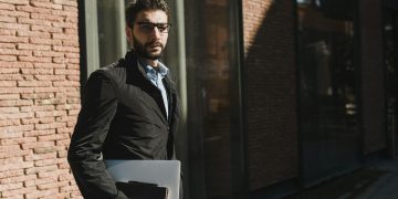 Stylish man in black jacket with glasses holding a laptop outside a glass building.