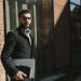 Stylish man in black jacket with glasses holding a laptop outside a glass building.
