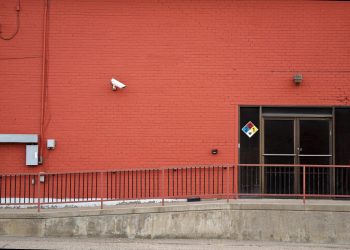 Urban red brick wall with security camera and entrance, featuring safety signage and railing.