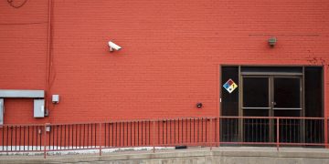 Urban red brick wall with security camera and entrance, featuring safety signage and railing.