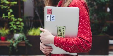 A young woman in a red polka dot shirt holds a laptop with stickers outdoors.