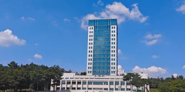 Tall office building under clear blue sky, surrounded by trees and grass lawn.