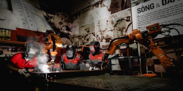 Three welders in protective gear working with robots in a high-tech industrial workshop.