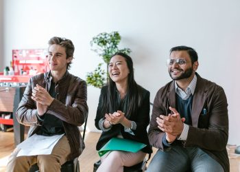 Three diverse team members clapping and smiling during an indoor meeting, showing positivity and teamwork.