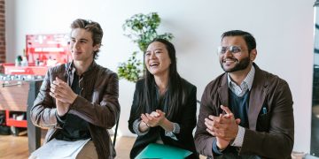 Three diverse team members clapping and smiling during an indoor meeting, showing positivity and teamwork.