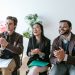 Three diverse team members clapping and smiling during an indoor meeting, showing positivity and teamwork.
