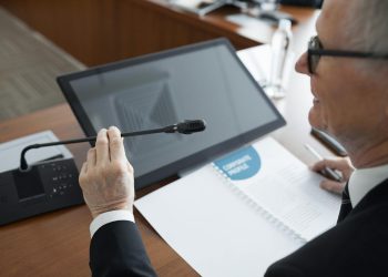 A senior businessman using a microphone during a conference meeting with documents and a monitor.