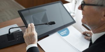 A senior businessman using a microphone during a conference meeting with documents and a monitor.