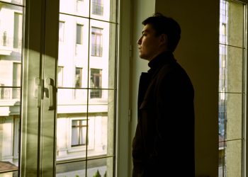 Contemplative man standing by a window indoors, gazing outside.