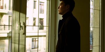 Contemplative man standing by a window indoors, gazing outside.