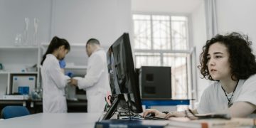 Focused woman working on a computer in a busy laboratory setting, showcasing teamwork and scientific research.