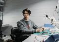 Asian man working with soldering tools and microscope on electronics in a lab setting.