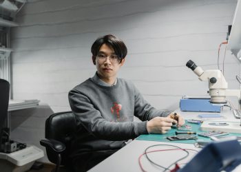 Asian man working with soldering tools and microscope on electronics in a lab setting.