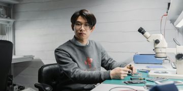 Asian man working with soldering tools and microscope on electronics in a lab setting.