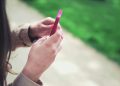 Woman holding a pink smartphone with both hands in a casual outdoor setting.