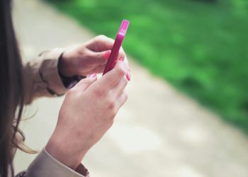 Woman holding a pink smartphone with both hands in a casual outdoor setting.