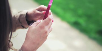Woman holding a pink smartphone with both hands in a casual outdoor setting.