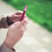 Woman holding a pink smartphone with both hands in a casual outdoor setting.
