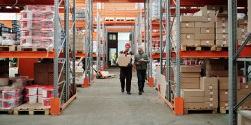 Two workers handle a package in a spacious warehouse surrounded by shelves stocked with boxes and products.