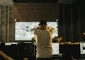 A man stands thoughtfully in a dimly lit control room with various screens.
