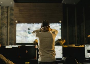 A man stands thoughtfully in a dimly lit control room with various screens.