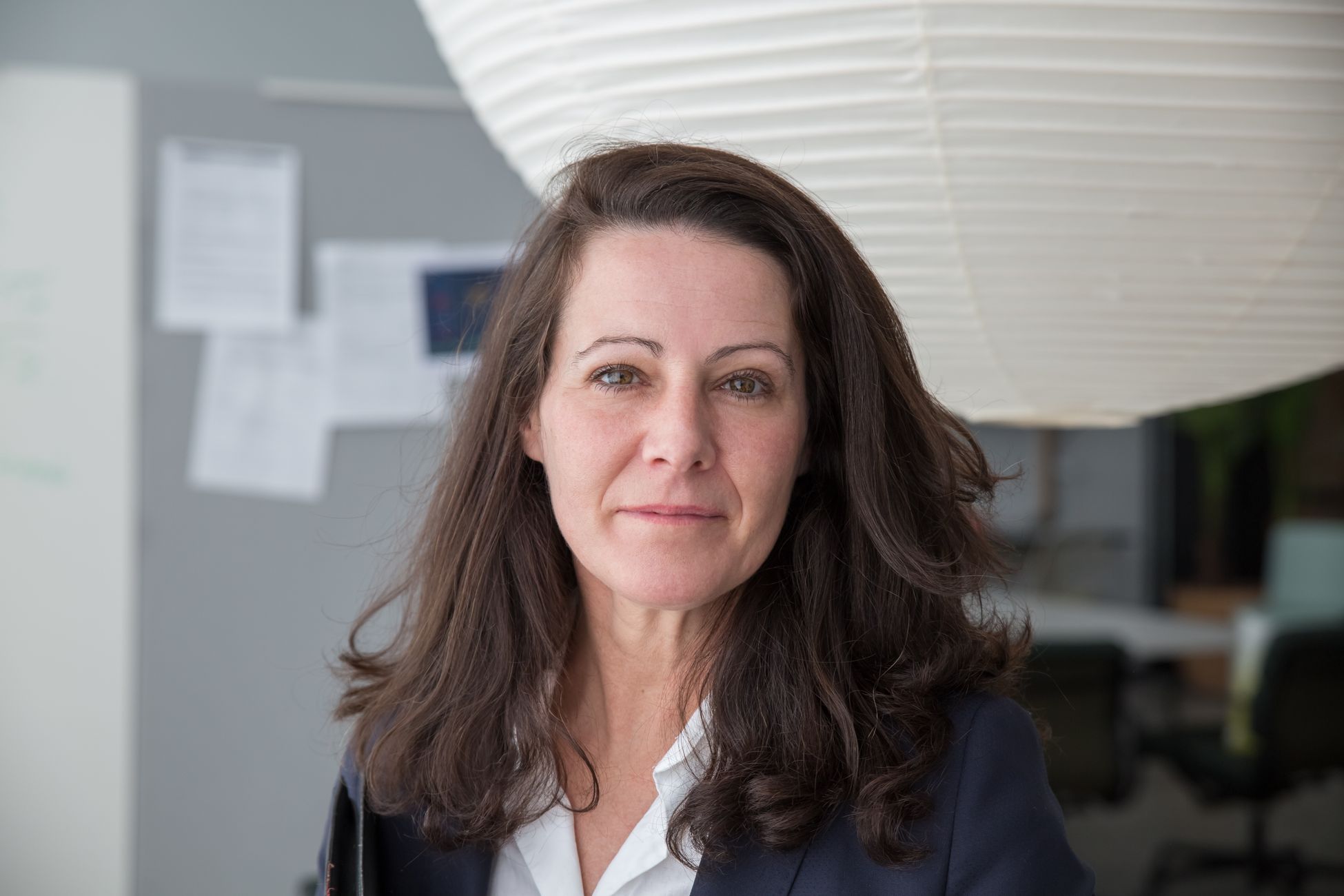 Portrait of a confident businesswoman in a modern office, wearing a blazer with a neutral expression.