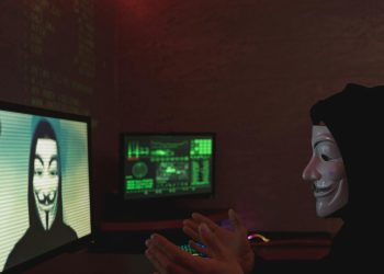 Masked individual in dark room interacting with computer screens displaying hacker imagery and code.