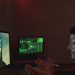 Masked individual in dark room interacting with computer screens displaying hacker imagery and code.