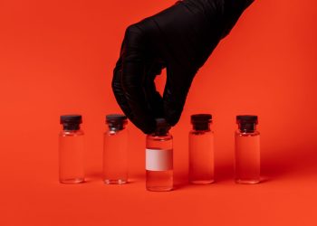 Person wearing black glove grabbing a vaccine vial against a bright red backdrop.