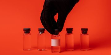 Person wearing black glove grabbing a vaccine vial against a bright red backdrop.