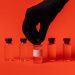 Person wearing black glove grabbing a vaccine vial against a bright red backdrop.