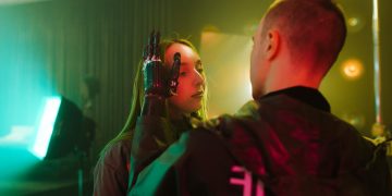 A man with a prosthetic hand gently touches a woman's face in a cyberpunk setting.