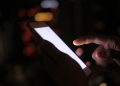 Close-up of a hand using a smartphone in the dark with blurred city lights in the background.