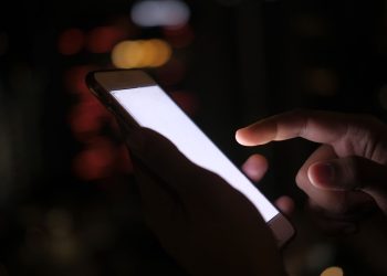 Close-up of a hand using a smartphone in the dark with blurred city lights in the background.