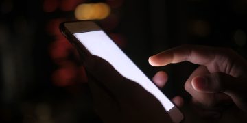 Close-up of a hand using a smartphone in the dark with blurred city lights in the background.